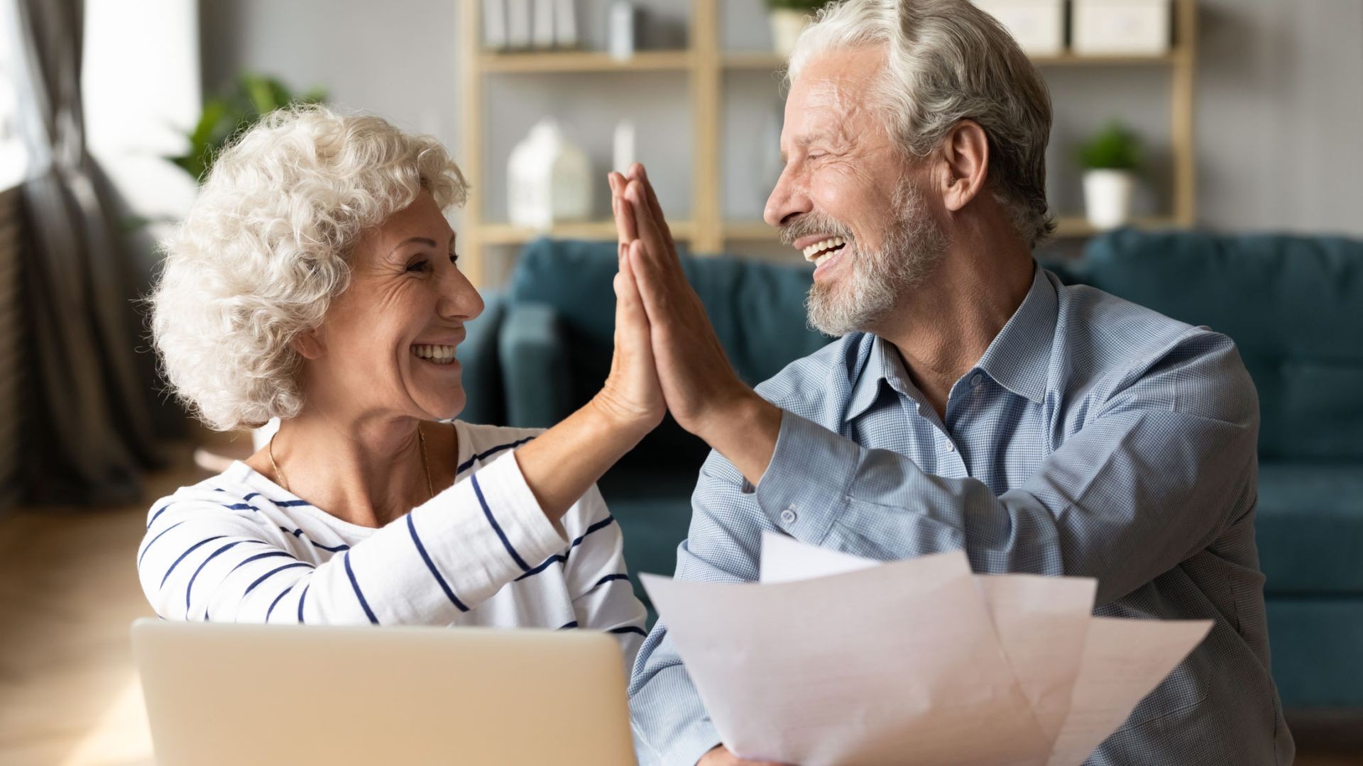 Happy elderly couple high-fiving each other while sitting at a table with a laptop and papers.